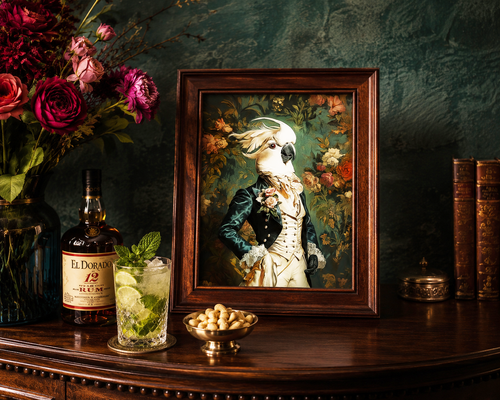 Framed portrait of a parrot in formal attire on a wooden table with a bottle of El Dorado rum, cocktail, and flowers.
